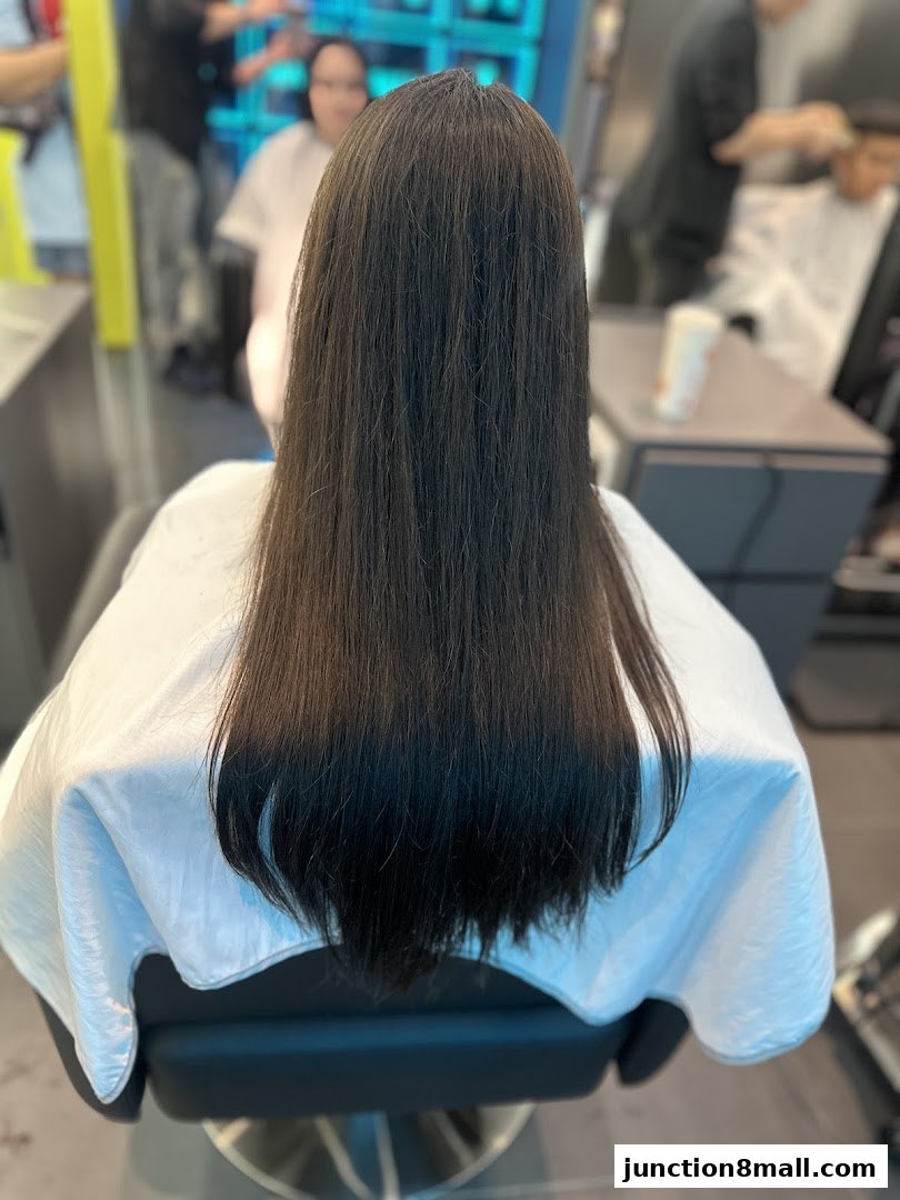 A person's long dark hair is styled in a hair salon, with stylists blurred in the background.