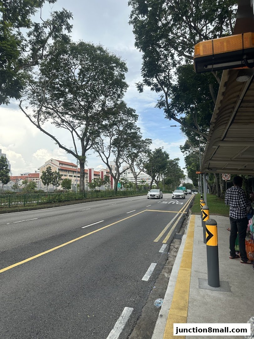 A street view with trees, a road, and a bus stop shelter with people waiting.