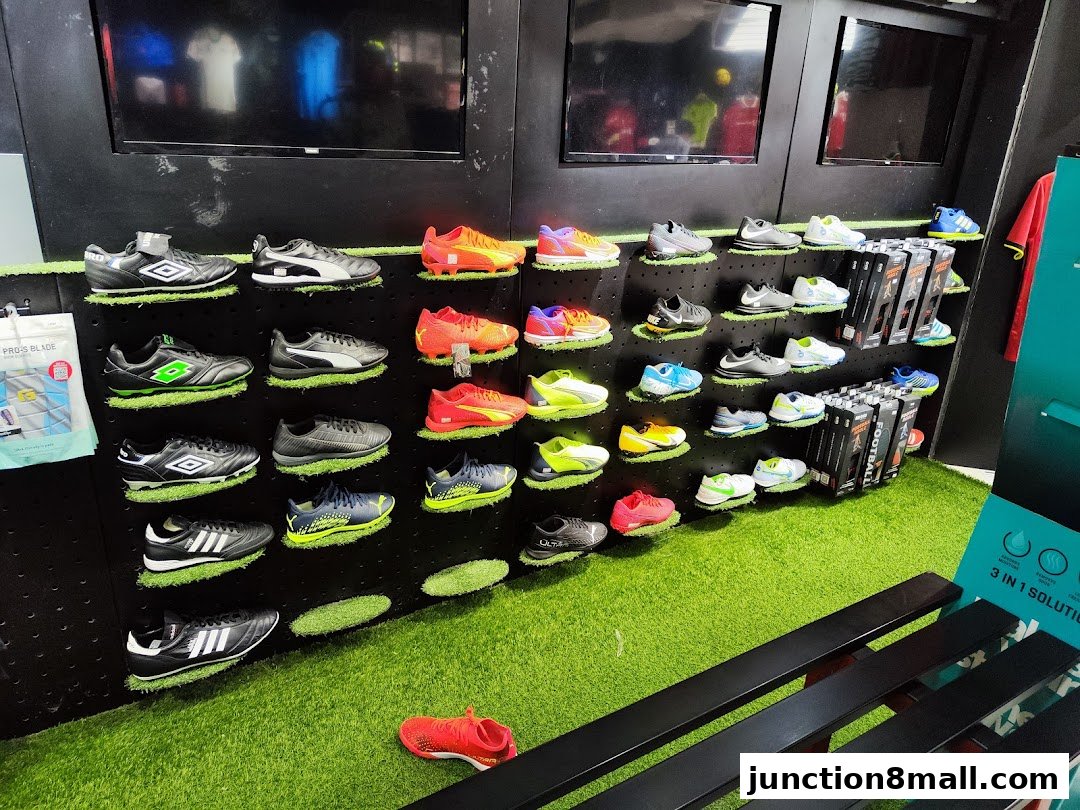 A display of various soccer cleats on shelves with artificial grass and screens in the background.