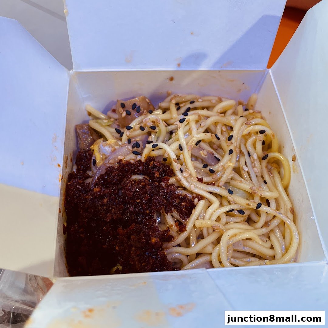 A close-up of a takeaway box filled with noodles, chili paste, and sesame seeds.