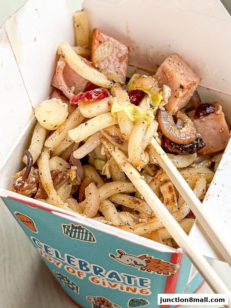 A close-up shot of a takeaway box filled with noodles, meat, and vegetables, with chopsticks resting on top.
