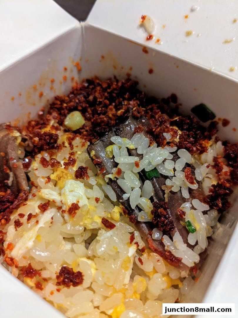 A close-up view of a takeaway box filled with rice, meat, and chili flakes.