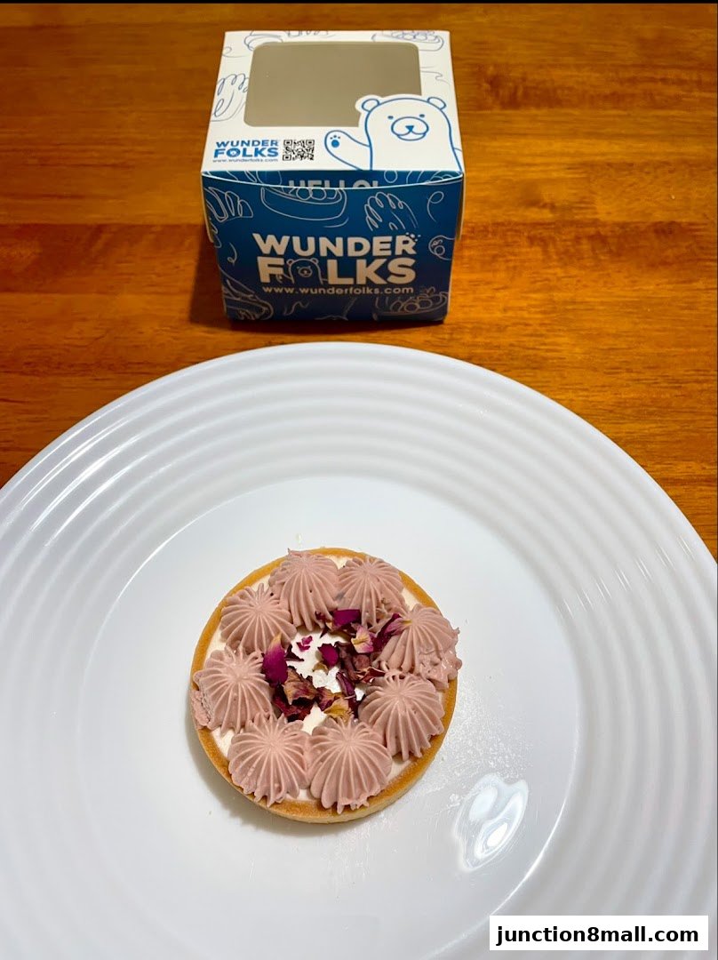 A beautifully decorated tart with pink frosting and rose petals on a white plate, next to a branded box.