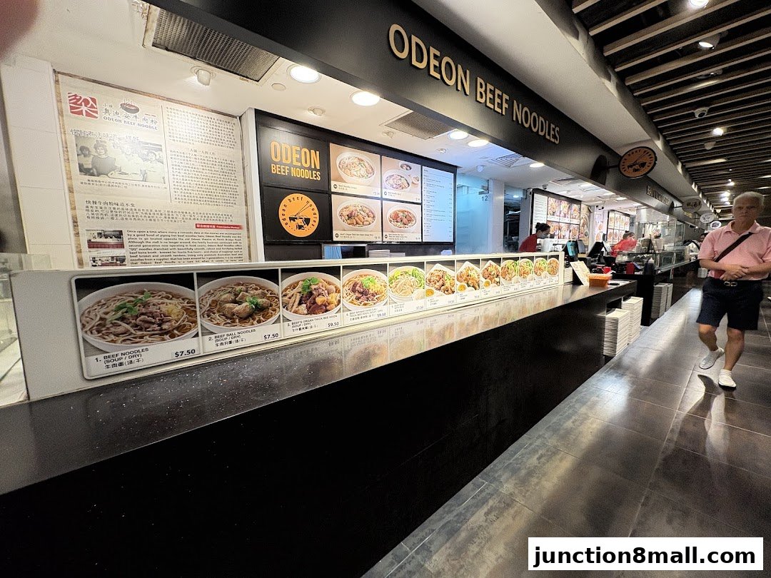 A food stall counter displaying various beef noodle dishes with prices and descriptions.