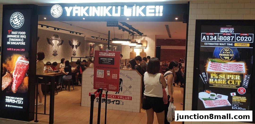 The entrance of Yakiniku Like, a Japanese BBQ restaurant, with customers inside and a digital display showing wait times and promotions.