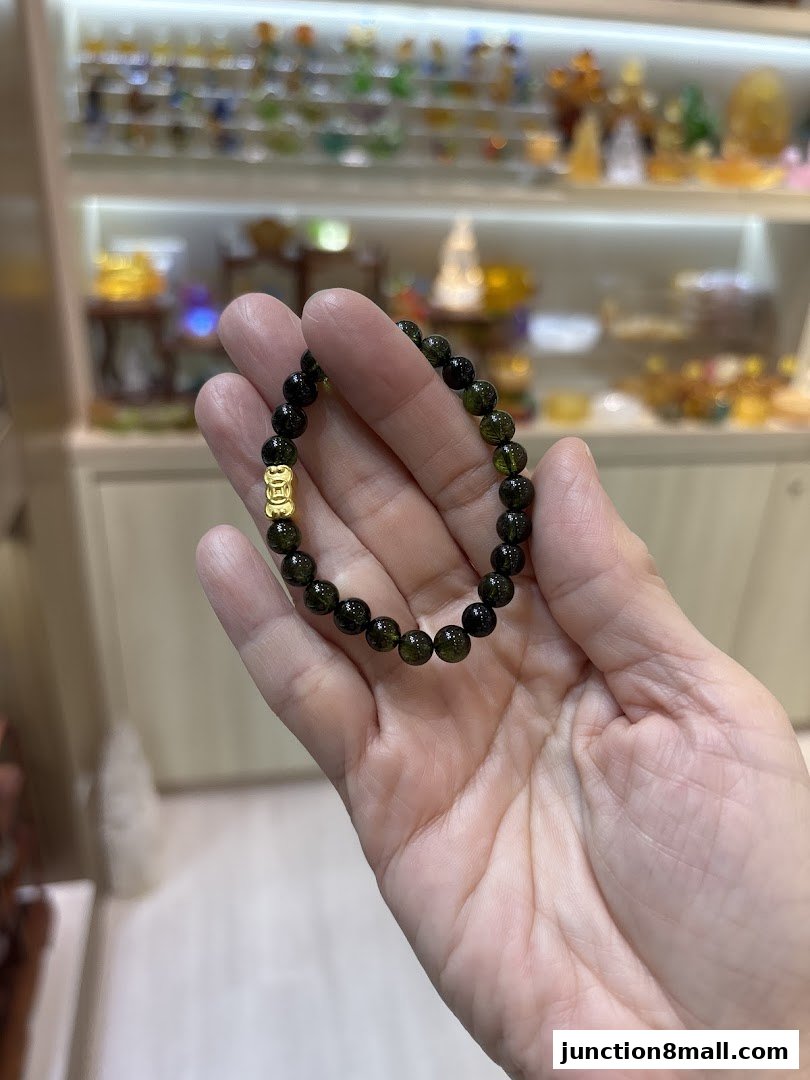 A hand holds a green beaded bracelet with a gold charm in front of a display of various items.