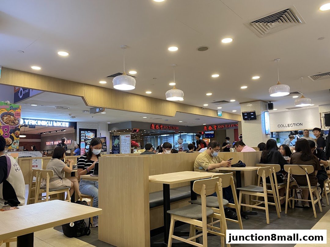 Diners seated at tables inside a brightly lit food court with various restaurant stalls visible.