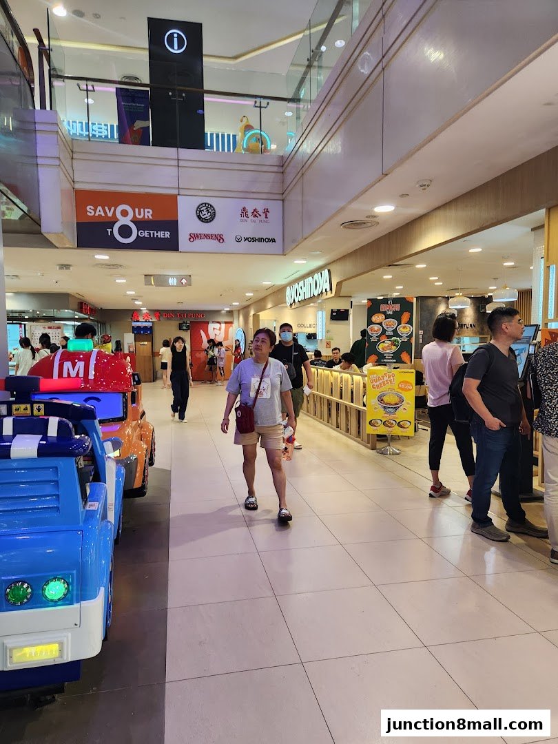 People walking in a mall hallway with various shop signs and a children's ride.